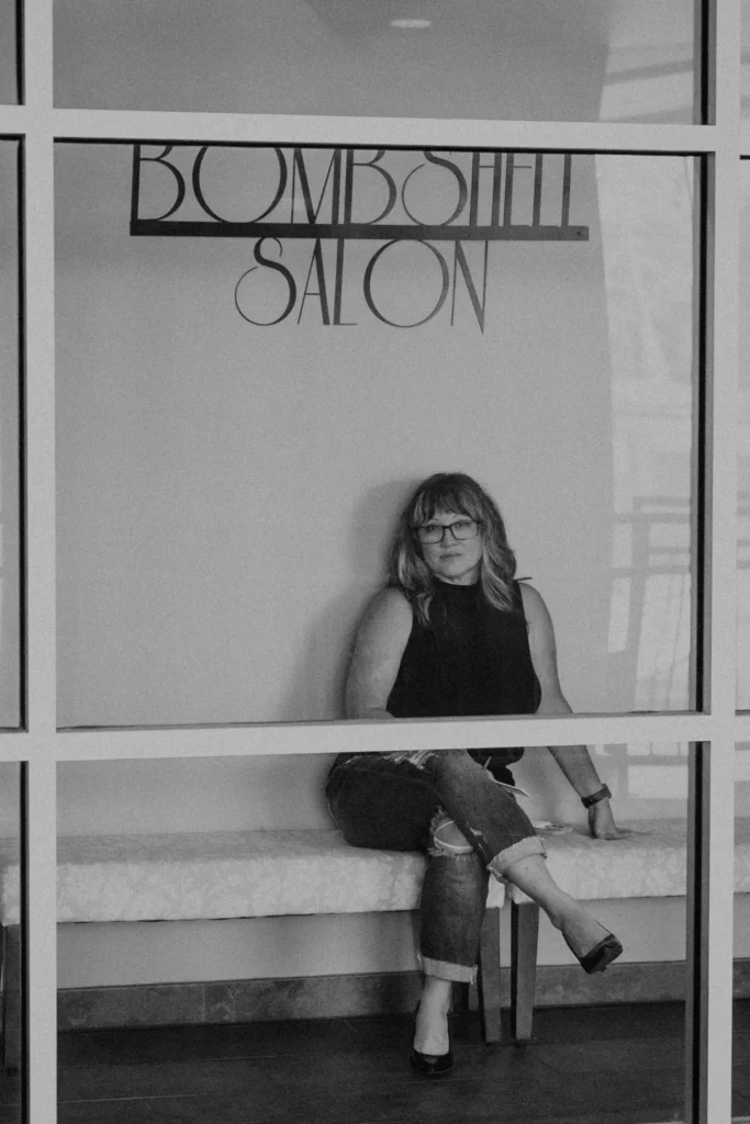 Diane Lichtas sitting in the hallway on a bench under the Bombshell Salon sign