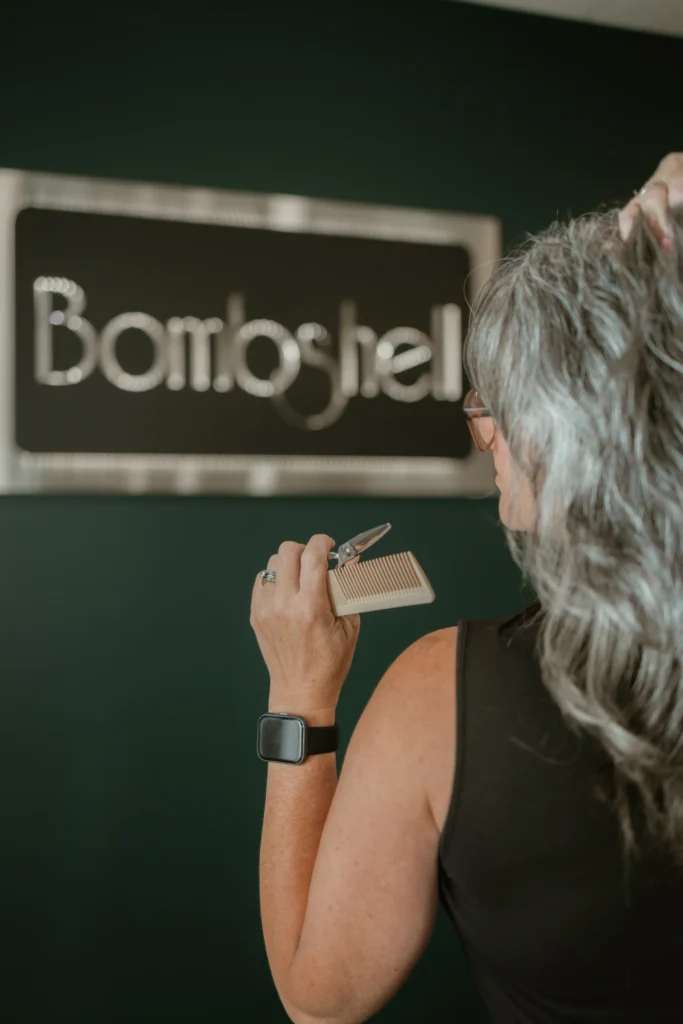 Diane Lichtas holds a large comb and shears while standing in front of a green wall that says "Bombshell".