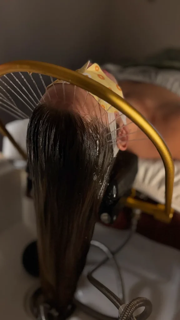 Person receiving hydrotherapy treatment under circular golden shower head with water cascading over wet dark hair.